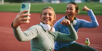 Two women sitting on a running track, smiling and flexing their arms while taking a selfie. One has headphones around her neck, and the other sports a compression wrap on her wrist.