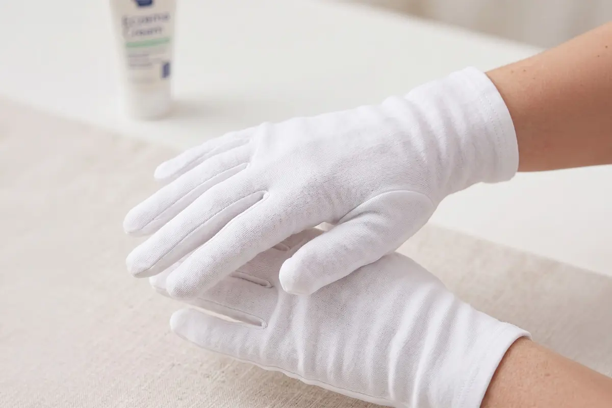 Two hands wearing white cotton gloves for dermatitis are placed on a light-colored surface, with a tube of cream in the background.