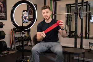 A man sits on a gym bench, holding a red resistance band while speaking to a smartphone camera mounted on a ring light in a fitness studio, sharing details about the gym’s ambassador program.