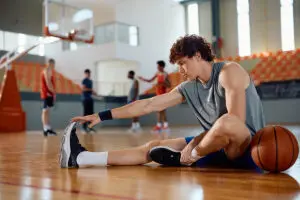 Young man stretches his leg while sitting on a basketball court, with a basketball by his side and other players blurred in the background—a moment that highlights the importance of high school athlete recovery tips to help manage fatigue in high school athletes.