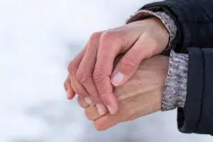 Two hands with signs of dry hands clasped together, wearing a black jacket with a grey knit cuff against a snowy background.