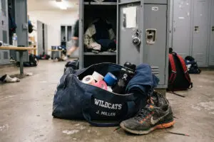 Open gym bag labeled Wildcats J. Miller sits on locker room floor, with athletic gear, athletic tape and wraps, and a shoe next to nearby lockers.