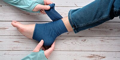 A person applies a blue compression wrap around another person's ankle, who is wearing jeans, on a wooden floor.