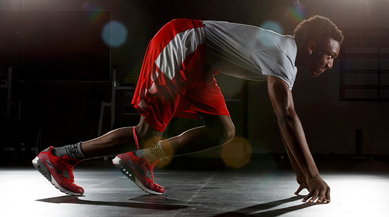 A man in athletic wear crouches in a sprinter’s starting position indoors, wearing a gray shirt, red shorts, red sneakers, and a compression wrap on his knee.