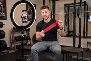 A man sits on a gym bench, holding a red resistance band while speaking to a smartphone camera mounted on a ring light in a fitness studio, sharing details about the gym’s ambassador program.