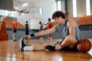 Young man stretches his leg while sitting on a basketball court, with a basketball by his side and other players blurred in the background—a moment that highlights the importance of high school athlete recovery tips to help manage fatigue in high school athletes.