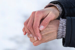 Two hands with signs of dry hands clasped together, wearing a black jacket with a grey knit cuff against a snowy background.
